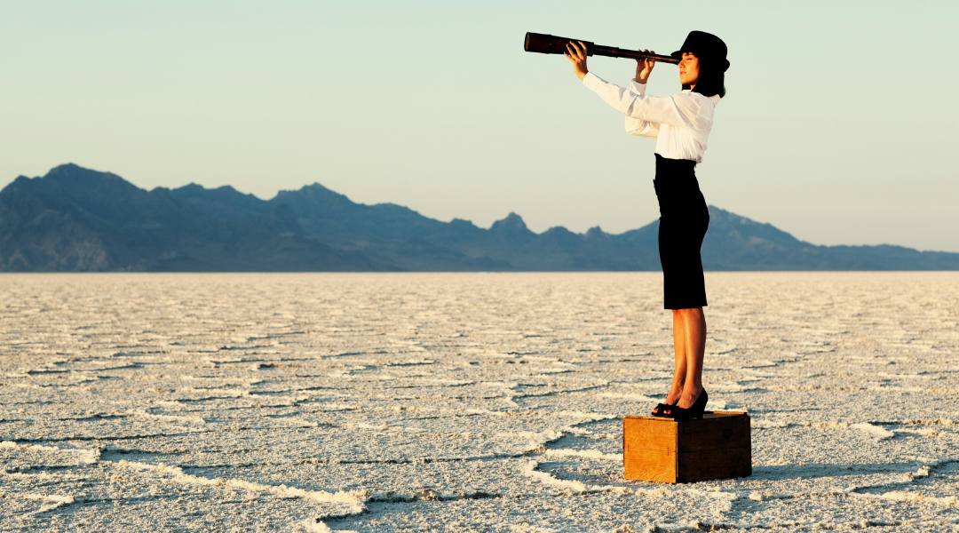 business woman standing on a crate in desert focusing her sights through a telescope 