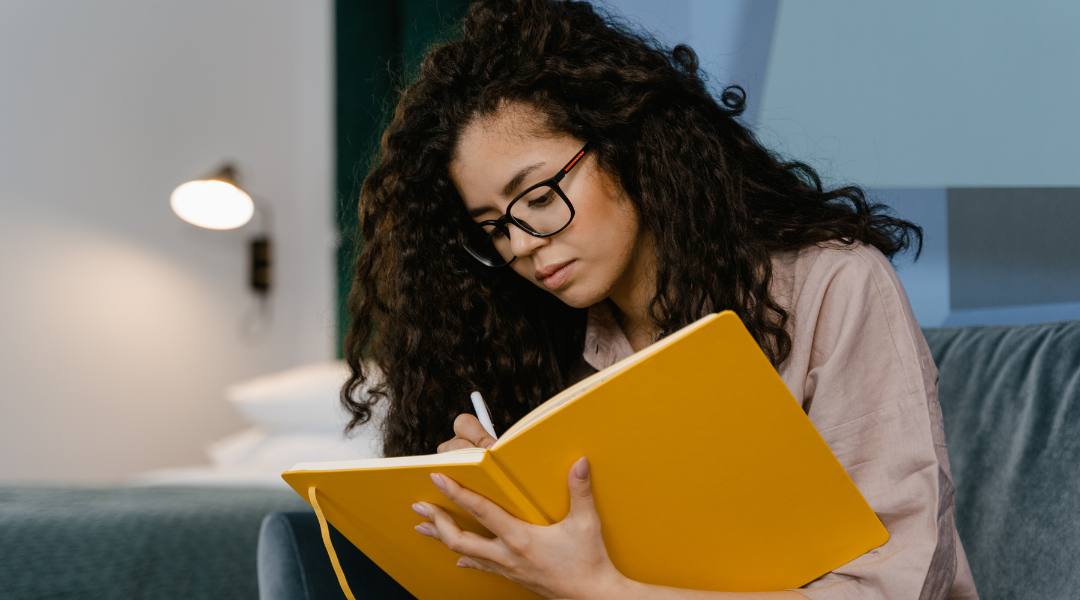 woman writing in a yellow leather journal thinking about how to keep your one-person business moving forward
