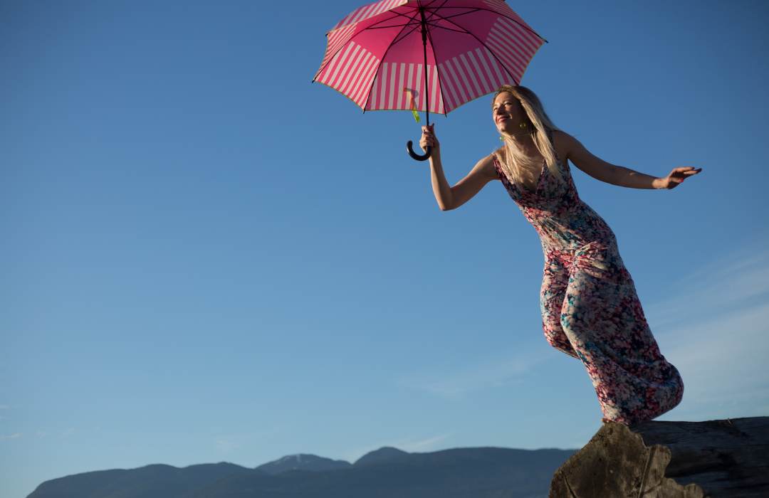 woman in flowing dress, leaning off a cliff, smiling and holding pink umbrella ready to embrace change